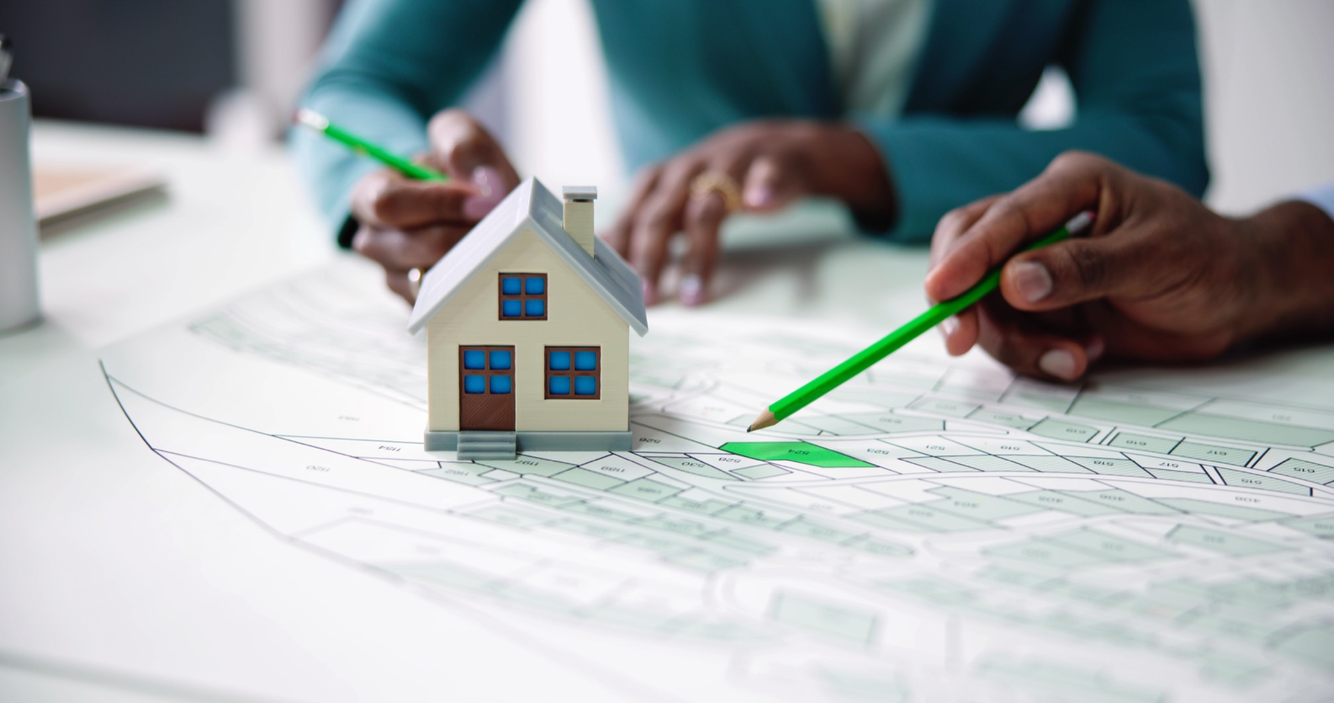 Ein Modellhaus steht auf einem Architektenplan. Im Hintergrund sind zwei Hände mit jeweils einem Stift zu sehen.